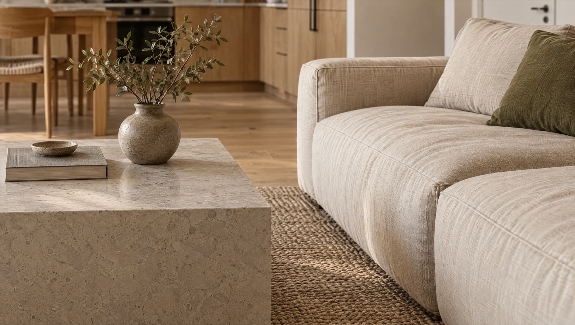 Close render across a stone coffee table and a linen sofa on a jute rug, the oak kitchen soft behind them.